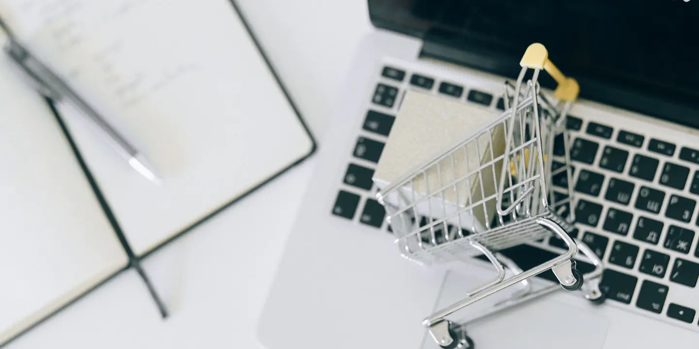 A miniature shopping cart holding a small box on a laptop keyboard, next to an open notebook and pen.
