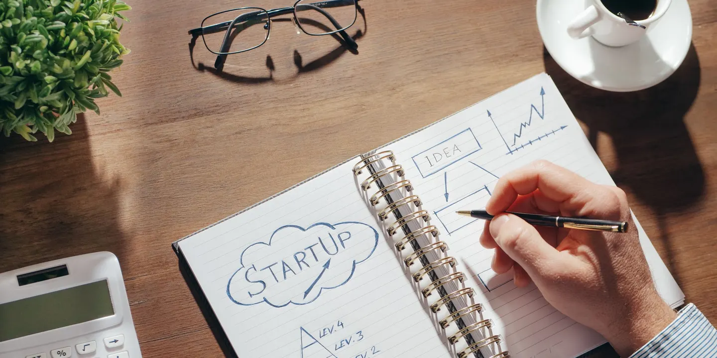 Seen from above, a hand writing in a spiral notebook with “Startup” drawn in a cloud and a small flow chart, next to a cup of black coffee, glasses, a calculator and a plant on a wooden desk.