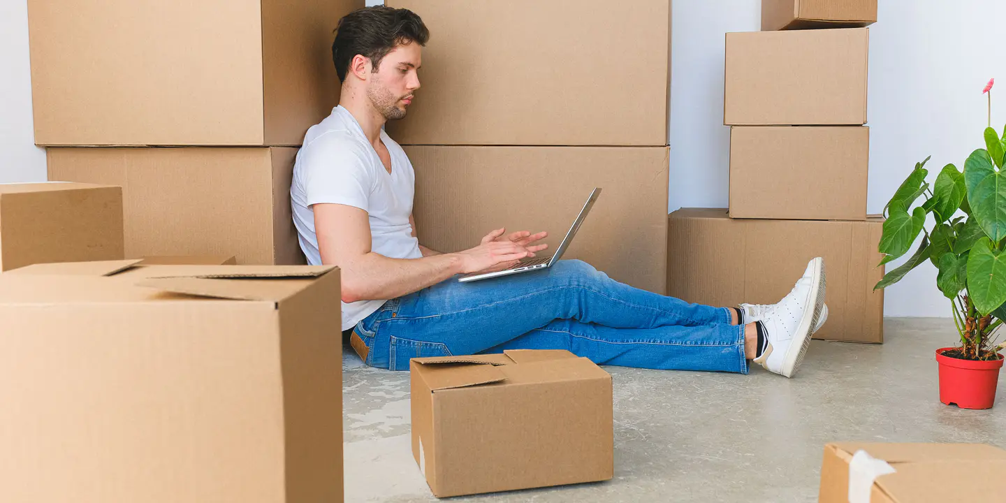 A man sitting on the floor typing on a laptop, surrounded by stacked cardboard moving boxes and a potted plant.
