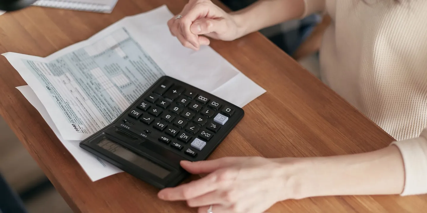 A person at a wooden table resting a finger on a large black calculator placed on printed forms, with a white mug in the foreground.
