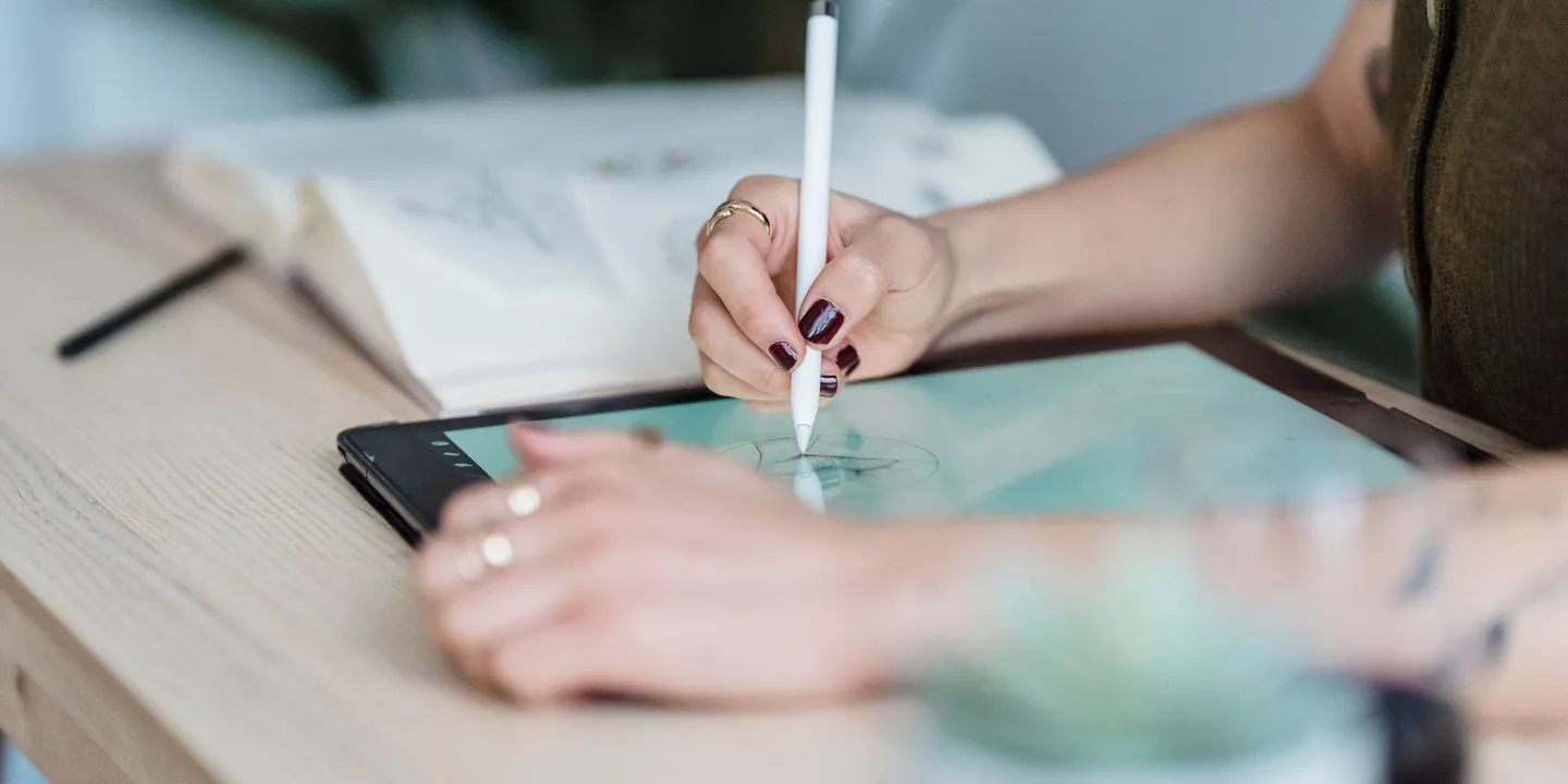 A woman with dark red nail polish sketching on a tablet with a white stylus at a light wooden desk, with an open sketchbook behind her hands.