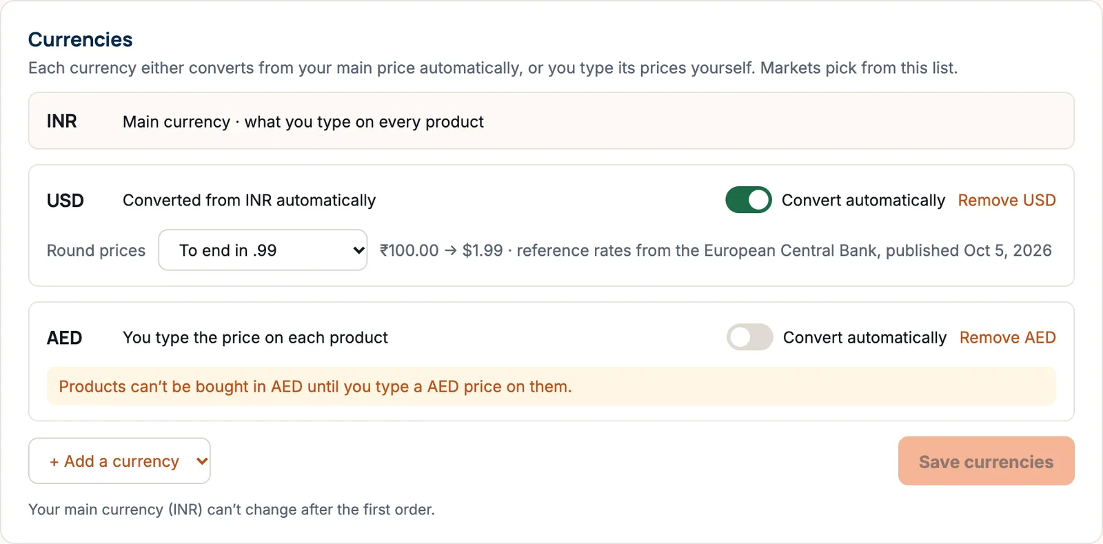 The Currencies card: INR as the main currency, USD converted from INR automatically with Round prices set to To end in .99 and an example of 100 rupees becoming 1.99 dollars, and AED with Convert automatically off and a note that products can’t be bought in AED until a price is typed.